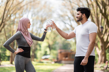 Wall Mural - Young woman in hijab and muslim man making cheers with bottles of water while standing at green summer park. Healthy active couple in sport clothes enjoying outdoors workout.