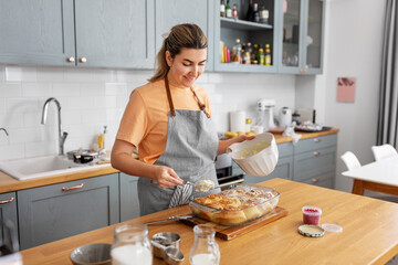 Wall Mural - culinary, bake and cooking food concept - happy smiling young woman adding buttercream topping to baked roll buns in baking dish on kitchen at home