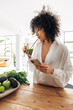 © Daniel - Young african american woman looking at her phone, browsing through social media while having a green juice for breakfast. Reusable bamboo straw. Big loft apartment. Vertical