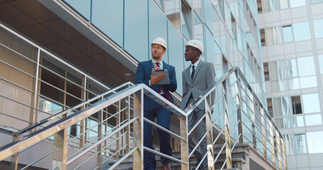 Diverse business partners in hardhat walking downstairs outside modern building and using digital tablet