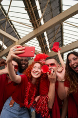 Vertical photo of a group of people taking a smiling selfie while cheering for their sports team from a stadium. Cheerful fans take a cell phone photo while celebrating at the stadium.