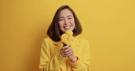 Wall Mural - Cheerful Asian woman tempting to eat appetizing ice cream licks lips looks at delicious frozen summer dessert wears formal jacket isolated over yellow background. Junk food breaking diet concept