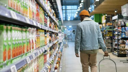 Wall Mural - Black man shopper in jacket wanders along long grocery shop rows looking around and holding plastic trolley close backside view