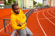 © shunevich - Young pensive handsome african american sportsman runner wearing yellow sportswear looking at camera, having rest after run training ar stadium, leaning on fence,