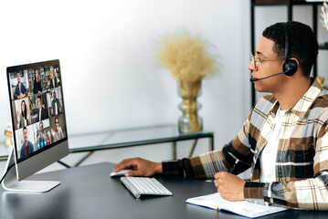 Wall Mural - Online consultation, video communication. Side view of a hispanic man with a headset communicating with business partners, discussing a project, conducts a consultation, multiethnic people on a screen