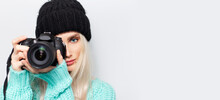 Portrait Of Blonde Girl Photographer Taking Photo On DSLR Camera, On White Background. Wearing Black Hat And Blue Sweater. Panoramic Photo With Copy Space.