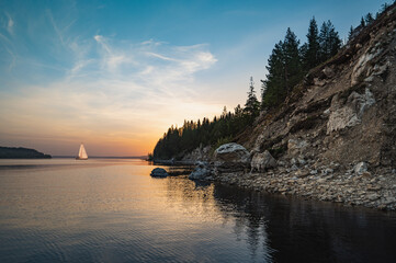 Wall Mural - Evening landscape, silhouette of a high, steep, rocky river bank at sunset reflection in the water, a sailing yacht sailing in the distance.
