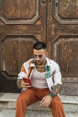 Wall Mural - Hot caucasian man with arm tattoos with a patterned shirt and brown pants looking at his phone