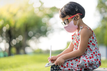 Wall Mural - Adorable little girl with protective face mask wearing wearing glasses sitting on park bench drawing on tablet.