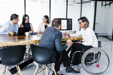 Wall Mural - Hispanic transgender businesswoman in wheelchair working on project with group of business people in office, in disability concept and disabled people