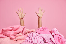 Female Hands Raised Up From Pile Of Unfolded Pink Clothes. Unrecognizable Cluttered Woman Poses Indoor. Monochrome Shot. Revision Of Clothing At Home. Disorder And Mass. Wardrobe Decluttering