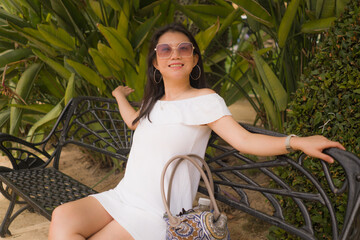 young beautiful and happy Asian woman on park bench - lifestyle portrait of Attractive Chinese girl in Summer dress resting during tourist walk tour