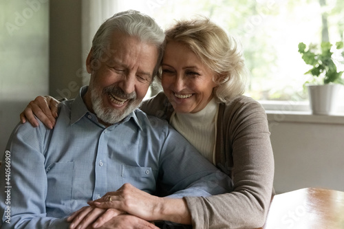 Smiling old middle-aged Caucasian man and woman sit hugging cuddling enjoy happy maturity life together at home. Excited optimistic mature couple spouses show love and care in family relationships.