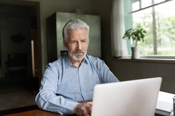 Wall Mural - Serious mature Caucasian man sit at desk at home office look at laptop screen work online on gadget. Pensive modern smart old 70s male use computer browse internet on web. Elderly technology concept.