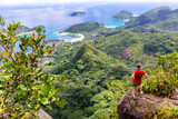 Young female traveller standing at the edge of the cliff at Morne Blanc View Point, overlooking Mahe Island coastline with lush tropical vegetation and crystal blue ocean, Seychelles.