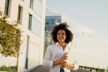 Wall Mural - A confident, curly-haired young woman of African-American appearance stands outside a business center in the city and smiles. Dressed in a white shirt. A confident person on the street. 
