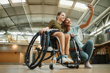 Low Angle Portrait Of Young Woman In Wheelchair Taking Selfie With Friend At Indoor Sports Court, Copy Space