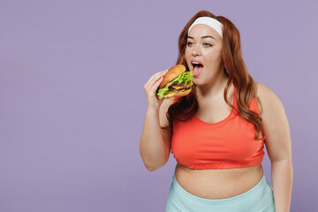 Young addicted chubby overweight plus size fat fit woman 20s wearing red top warm up training hold eating junk fast food biting burger isolated on purple background home gym. Workout sport concept