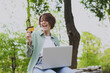 Young overjoyed fun woman in green jacket sit on bench in spring park outdoors rest use laptop pc computer hold credit bank card do online shopping order booking tour. People urban lifestyle concept.