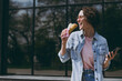 © ViDi Studio - Young hungry woman in jeans clothes pink eyeglasses stroll near glass building walk outdoors use fastfood hamburger sandwich have lunch use mobile cell phone lok aside People urban lifestyle concept.