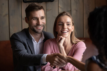 Happy beautiful young caucasian family couple shaking hands with professional realtor, broker or saleswoman, starting meeting in modern cafe. Smiling clients making agreement with real estate agent.