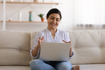 Millennial Indian woman sit on sofa talk speak on video call on computer with client customer. Young mixed race female have webcam digital virtual online conference on laptop. Communication concept.