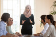 © fizkes - Smiling Asian muslim team leader talking to diverse employees at briefing, businesswoman wearing hijab speaking at corporate meeting, training or teaching staff, explaining project strategy