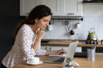 Wall Mural - Confident smiling businesswoman using laptop in kitchen, working on project online at home, focused young woman looking at computer screen, writing taking notes, student watching webinar, studying