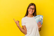 © Asier - Young caucasian woman holding bills isolated on yellow background smiling and pointing aside, showing something at blank space.