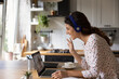 © fizkes - Side view excited woman in wireless headphones chatting online, waving hand at webcam, using laptop in kitchen, happy young female making video call at home, talking with colleague or friend