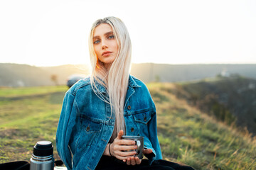 Portrait of young blonde girl in denim jacket holding steel mug on background of sunset.