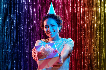 birthday, celebration and nightlife concept - happy smiling young african american woman in party cap with gift box over ultraviolet neon lights on rainbow foil curtain background