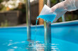 Hand holding and pouring algicide blue liquid from glass into water of swimming pool. Water purification and prevention of appearance of algae and microorganisms.