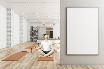 Young man in lotus pose sitting in modern yoga gym interior with empty poster on wall, equipment and daylight. Health and sport concept. Mock up.