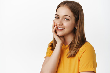 Wall Mural - Portrait of beautiful teen girl, touch skin without acne, no make up, touch cheek gently and smiling tender at camera, standing in yellow t-shirt against white background