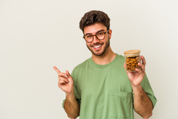 Young caucasian man holding an almond jar isolated on white background smiling and pointing aside, showing something at blank space.