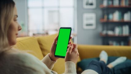 Poster - Beautiful Authentic Female Using a Smartphone with Green Screen Mock Up Display at Home Living Room while Lying on a Couch Sofa. She's Browsing the Internet and Checking Videos on Social Networks.