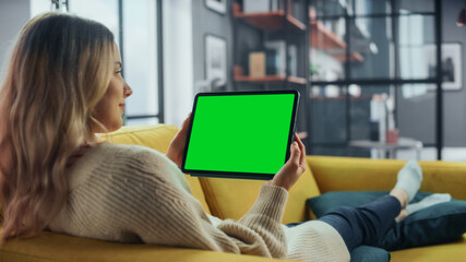 Aufkleber - Beautiful Authentic Female Using Tablet Computer with Green Screen Mock Up Display at Home Living Room while Lying on a Couch Sofa. She's Browsing the Internet and Checking Videos on Social Networks.