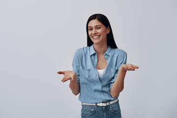 Wall Mural - Attractive young smiling woman looking at camera and gesturing while standing against grey background