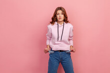 Portrait Of Upset Poor Student Girl In Hoodie And Jeans Turning Out Empty Pockets, Showing No Money Gesture, Worried About Debts. Indoor Studio Shot Isolated On Pink Background