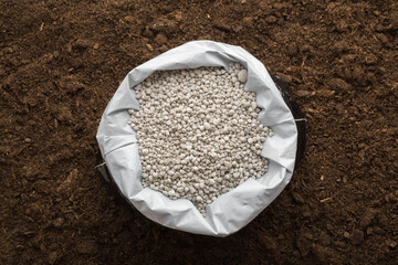 Opened plastic bag with gray complex fertiliser granules on dark soil background. Closeup. Product for root feeding of vegetables, flowers and plants. Top down view.