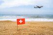 © flowertiare - Flag of Switzerland in the form of a toothpick in the sand of beach opposite sea wave with landing airplane. Travel concept