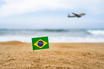 Flag of Brazil in the form of a toothpick in the sand of beach opposite sea wave with landing airplane. Travel concept