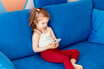 cute little toddler girl wear casual clothes sitting on blue sofa and watching cartoon on smartphone