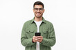 © Damir Khabirov - Young modern man in casual green shirt and glasses, holding smart phone in hands, looking at camera with smile, isolated on gray background