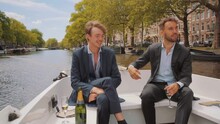 Caucasian Adult Men Dressed In Suit Navigating On The Canal Of Amsterdam In The Netherlands, On A Sunny Day. Luxury Tourism, Leisure Travel