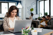 © DC Studio - Close up of satisfied businesswoman answering email typing on laptop smiling sitting at desk in busy start up office while diverse team analyzes statistics data. Multiethnic working at new project