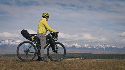 The man travel on mixed terrain cycle touring with bikepacking. The traveler journey with bicycle bags. Sport tourism bikepacking, bike, sportswear in green black colors. Mountain snow capped.