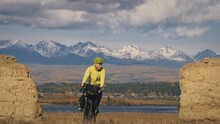 The Man Travel On Mixed Terrain Cycle Touring With Bikepacking. The Traveler Journey With Bicycle Bags. Sport Bikepacking, Bike, Sportswear In Green Black Colors. Mountain Snow Capped, Stone Arch.