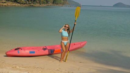 Pretty young sportswoman with sunglasses and swimsuit holds paddle posing near pink plastic canoe on sand beach at resort. Traveling to tropical countries. Attractive sexy girl posing near kayak.